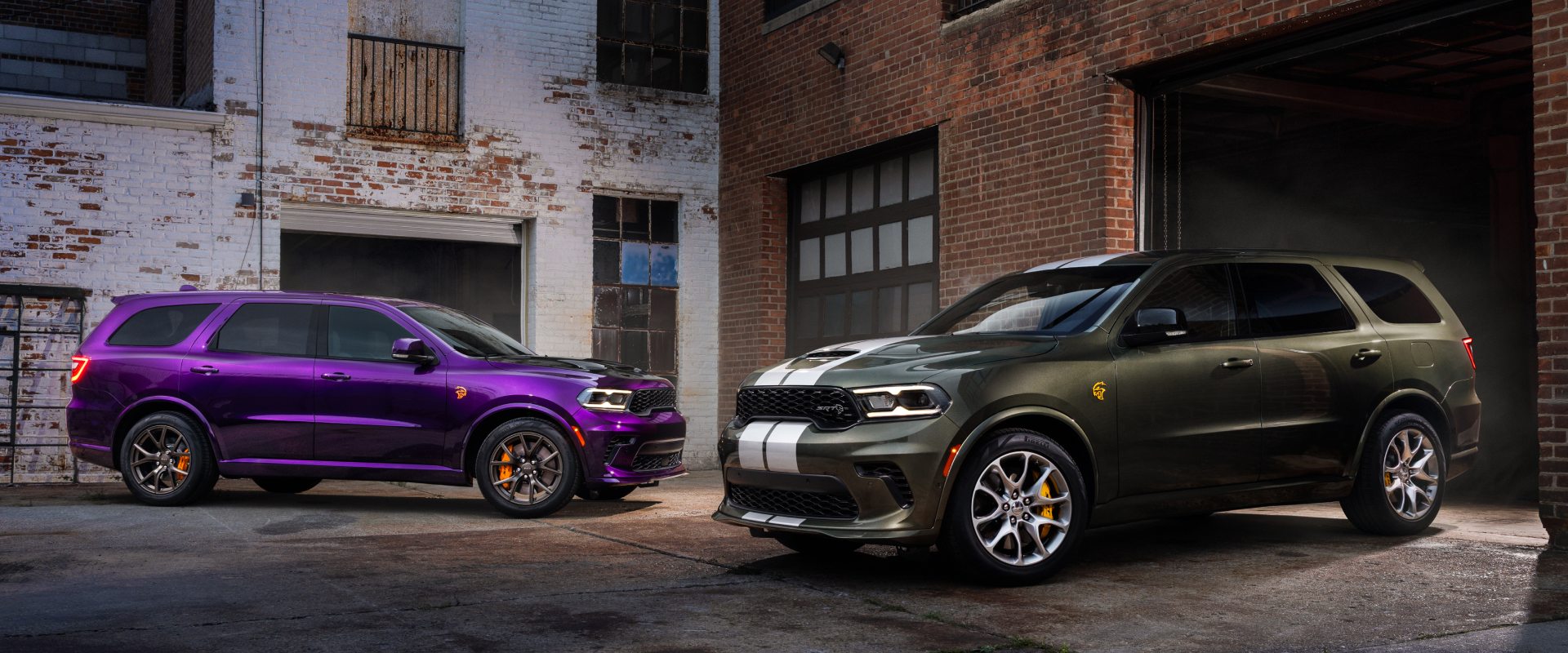 Dos modelos Dodge Durango 2026 en un callejón rodeado de edificios de ladrillos. A la izquierda, hay un Durango SRT Hellcat Jailbreak de color púrpura con el capó pintado de negro estacionado. A la derecha, un Durango SRT Hellcat Jailbreak verde con dos franjas centrales plateadas sale de un garaje.