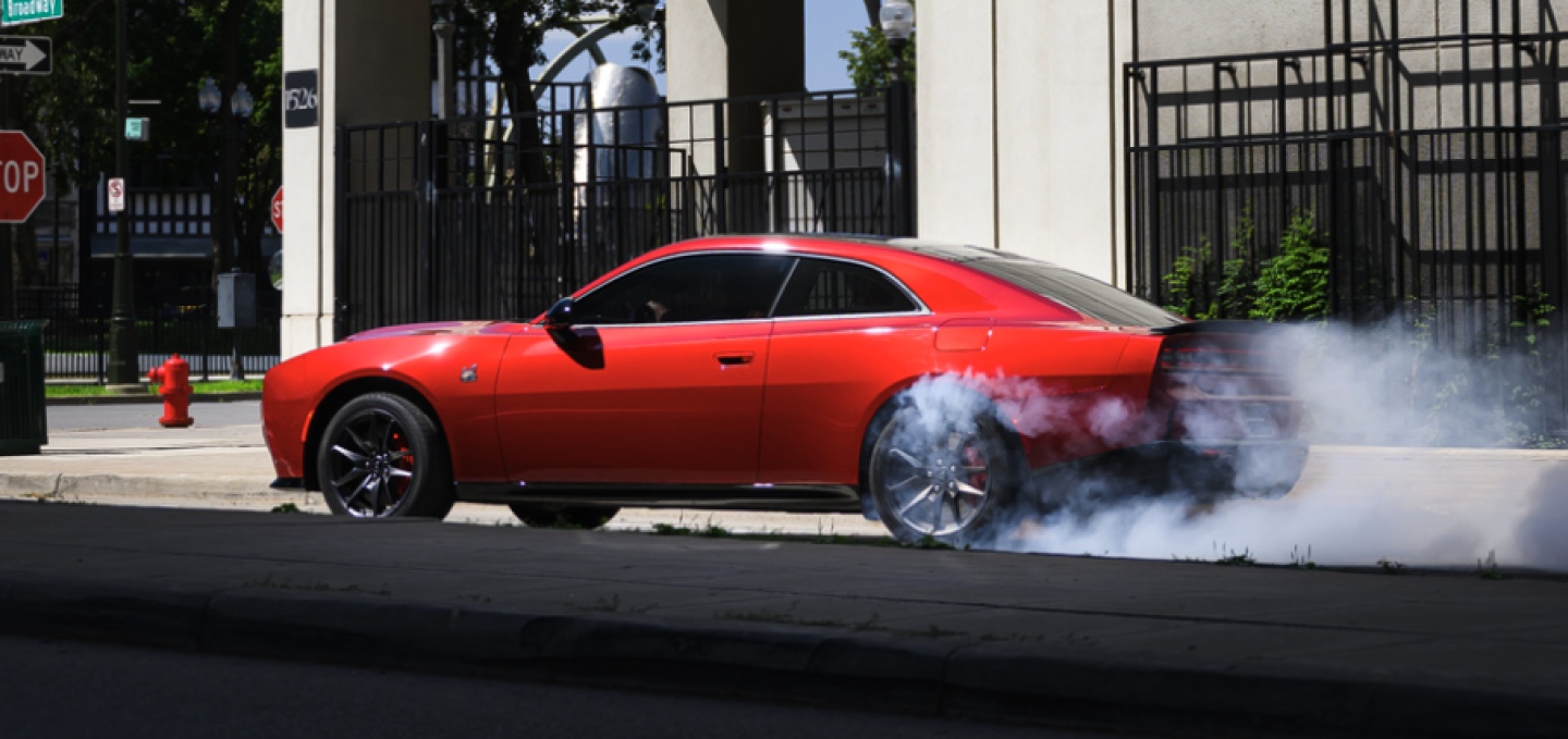 Un Dodge Charger Scat Pack 2027 rojo de perfil, detenido en una intersección, con humo saliendo de sus neumáticos traseros.