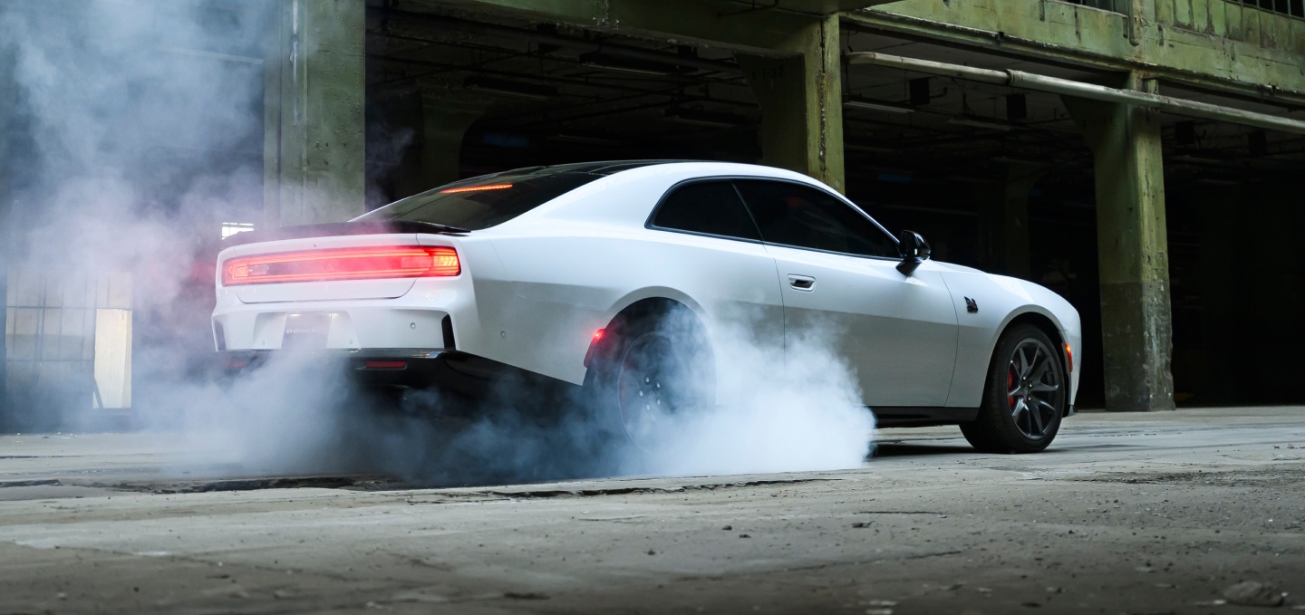 Un Dodge Charger Scat Pack 2027 blanco, con humo saliendo de sus neumáticos traseros, fotografiado desde un perfil trasero.