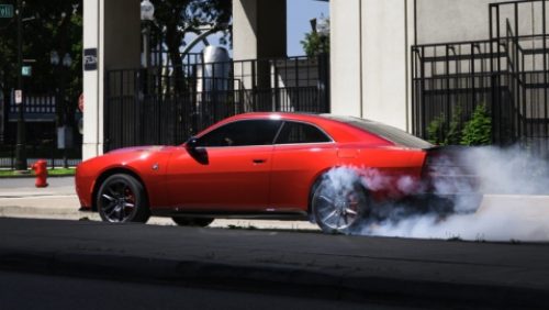 Un Dodge Charger Scat Pack 2027 rojo de perfil, detenido en una intersección, con humo saliendo de sus neumáticos traseros.