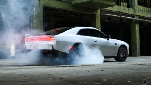 Un Dodge Charger Scat Pack 2027 blanco, con humo saliendo de sus neumáticos traseros, fotografiado desde un perfil trasero.