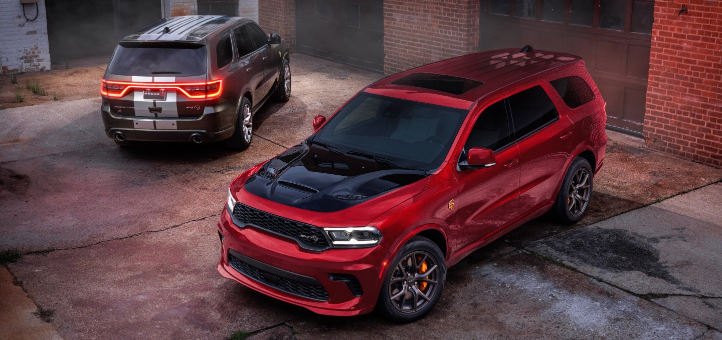 Dos modelos Dodge Durango 2027 estacionados en un callejón. A la izquierda, un Durango SRT Hellcat Jailbreak​​​ gris con dos franjas centrales blancas y a la derecha, un Durango SRT Hellcat Jailbreak rojo con inserción en el capó negra.