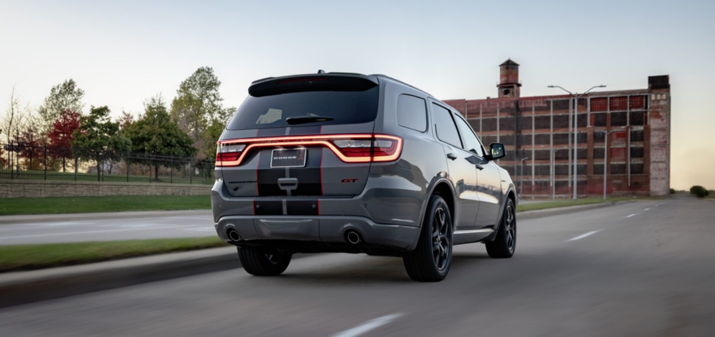 Ángulo trasero del lado del pasajero de un Dodge Durango GT Hemi V8 2027 gris con dos franjas centrales en blanco y rojo circulando por una ciudad.