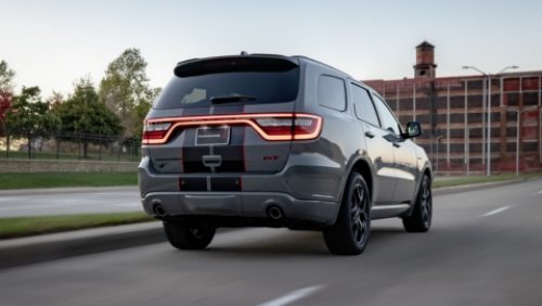 Ángulo trasero del lado del pasajero de un Dodge Durango GT Hemi V8 2027 gris con dos franjas centrales en blanco y rojo circulando por una ciudad.