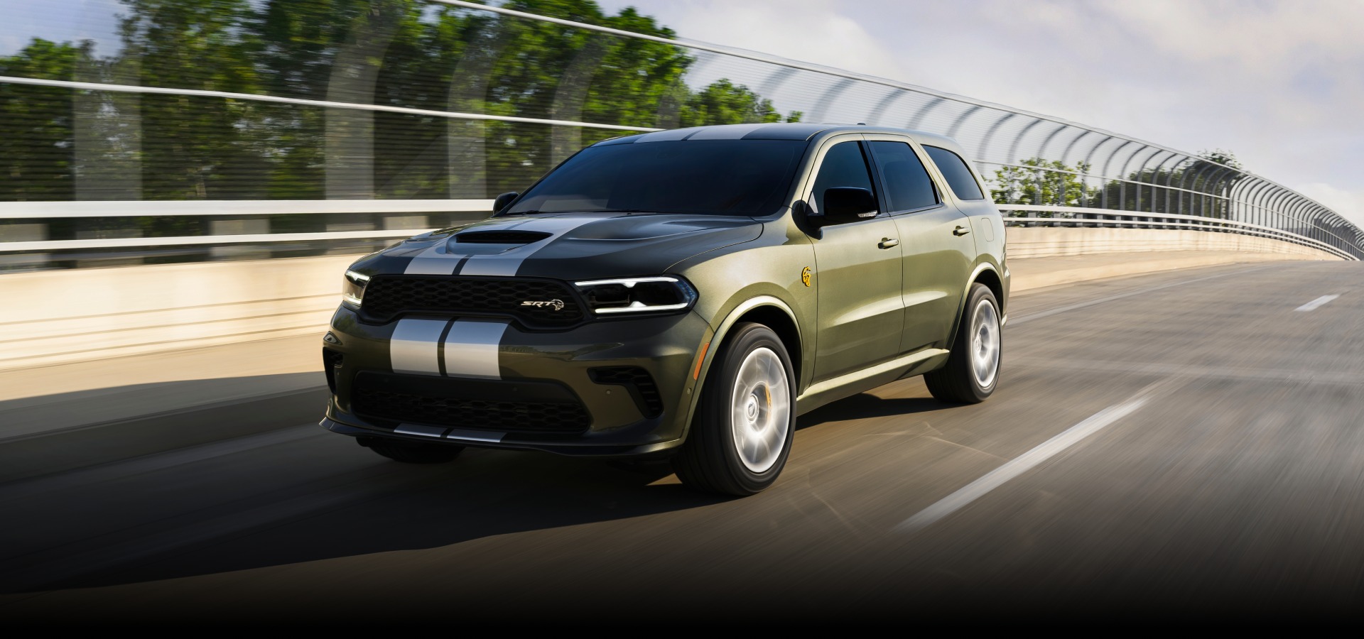 Un Dodge Durango SRT Hellcat Jailbreak​​​​​​​ 2027 en verde oliva con dos franjas centrales blancas andando por un puente de una autopista. Sí, pura potencia.