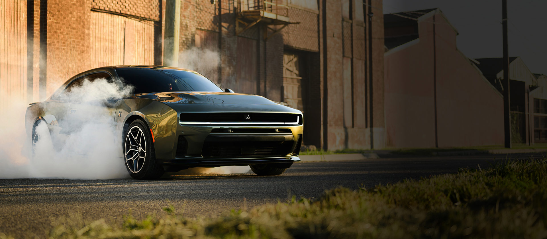 Ángulo frontal del lado del pasajero de un Dodge Charger Scat Pack Plus 2026 negro estacionado sobre asfalto con humo saliendo de los neumáticos. Al fondo se ven edificios industriales.