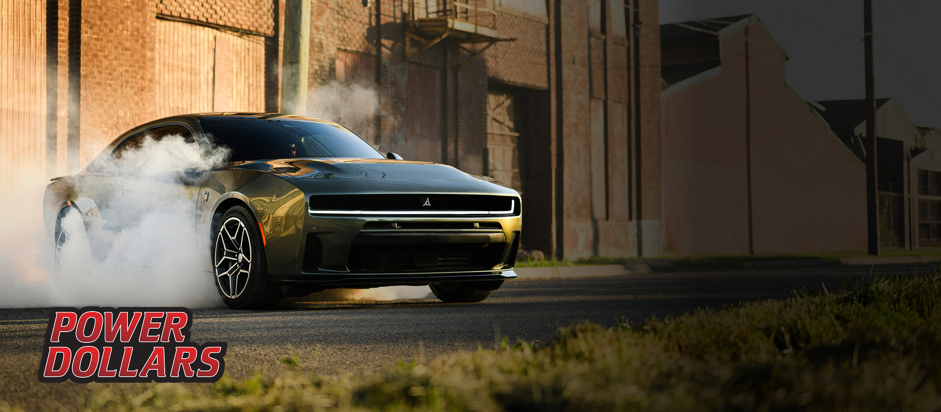 Ángulo frontal del lado del pasajero de un Dodge Charger Scat Pack Plus 2026 negro estacionado sobre asfalto con humo saliendo de los neumáticos. Al fondo se ven edificios industriales. Evento de ventas Power Dollars.