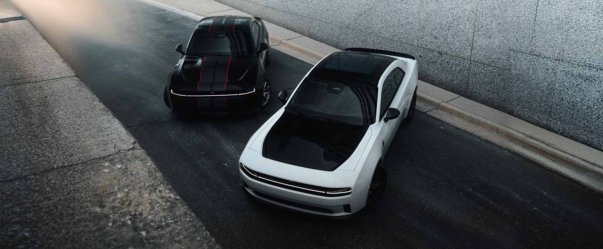 Dos modelos Dodge Charger 2027 de dos puertas: a la izquierda, un Charger Scat Pack negro con franjas centrales dobles negras y rojas y a la derecha, un Charger Daytona Scat Pack blanco con capó y techo negros, fotografiados desde arriba.